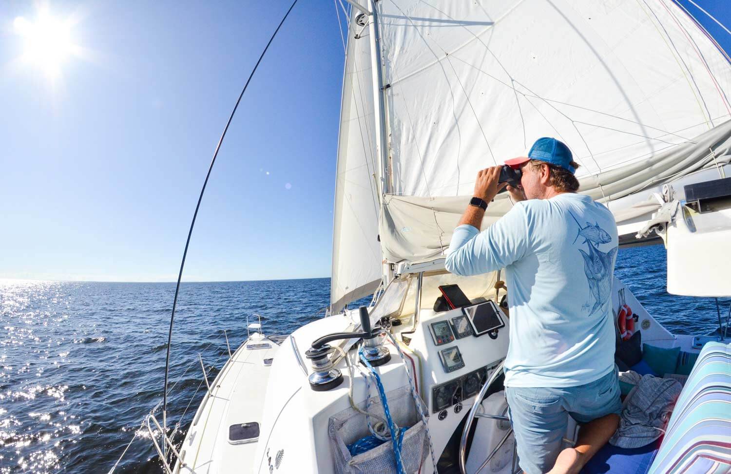 Captain at helm of a sailing catamaran looking through binoculars at horizon.