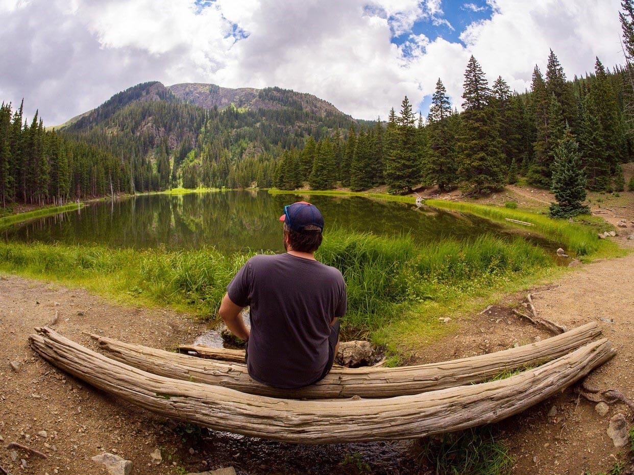 man sitting on log in front of mountain lake