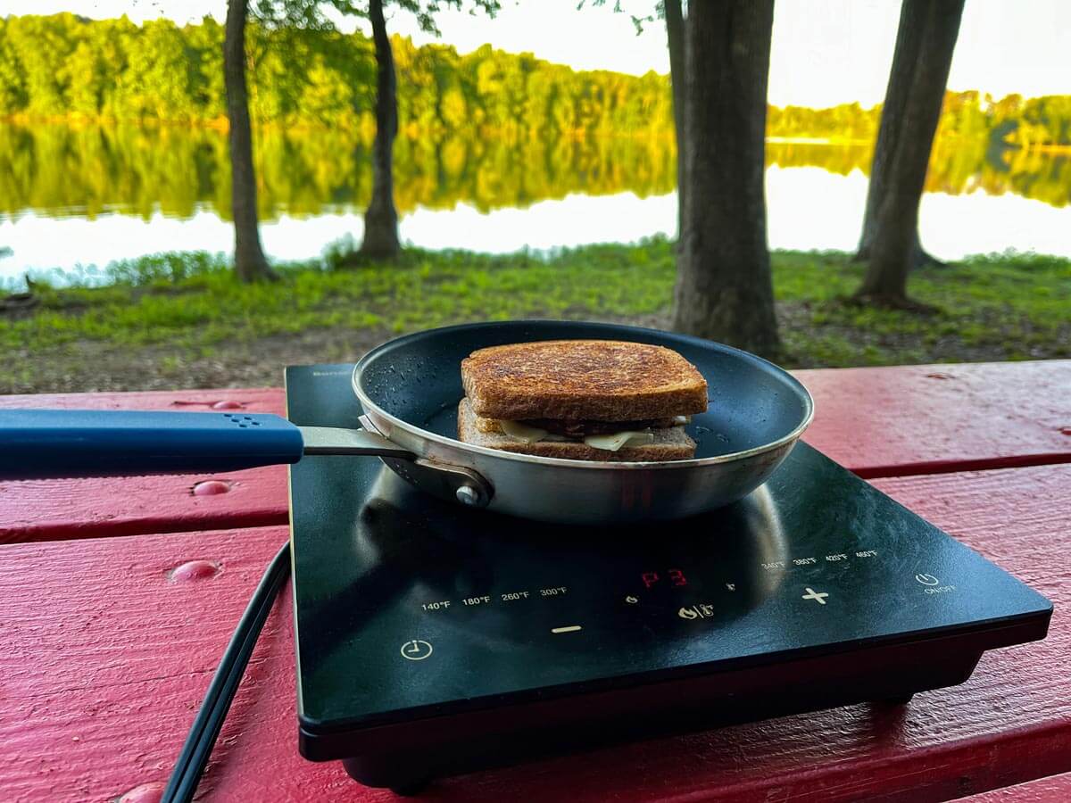 Small pan on induction plate with sandwich cooking outside at campground with lake in the background.