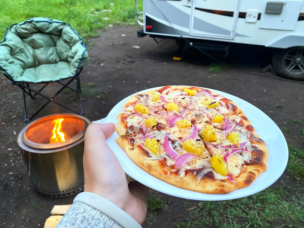 woman holding pizza on plate next to camping chair and campfire