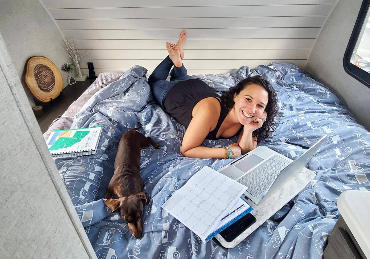 Woman laying on a bed with computer, paper, and writing materials planning a route for RVing the USA.