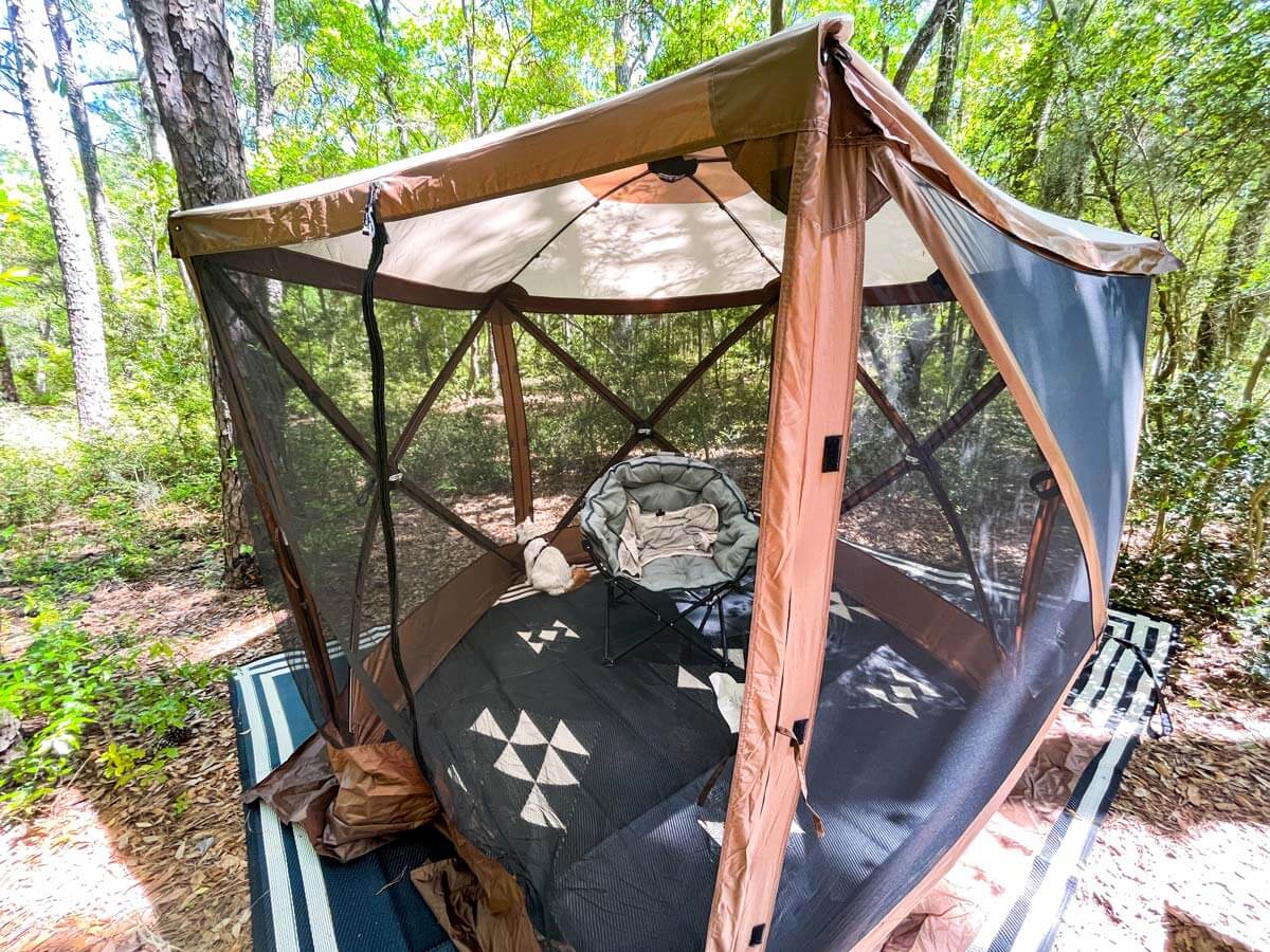 Clam pop up gazebo setup at a campsite in the woods with outdoor rug and camping chair inside.