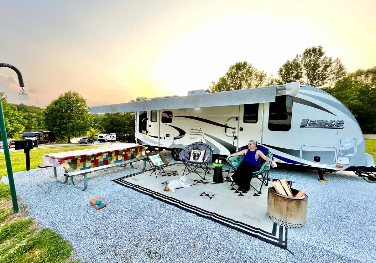 Woman sitting at RV campsite setup with a fire pit, multiple chairs, lanterns, a picnic table and more.