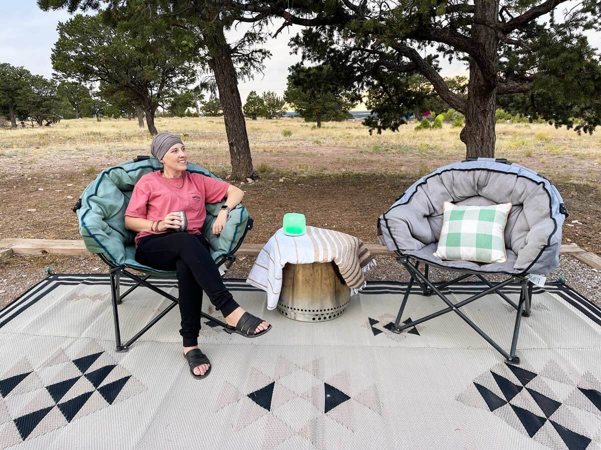 Woman sitting in a camping chair, with additional camping chair, outdoor rug, and solo stove as a table.