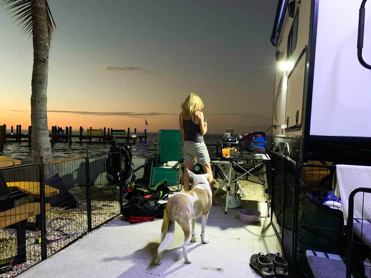 Dog and woman inside pet fence outside an RV at sunset by the water.