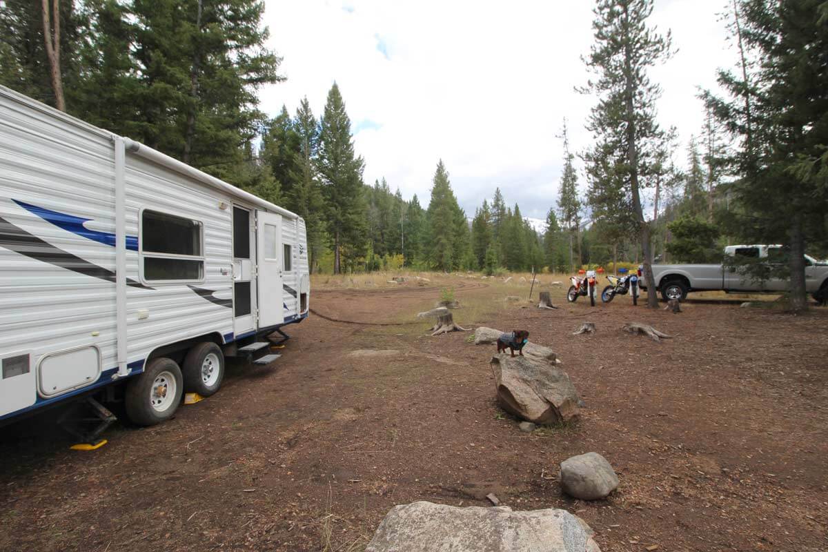 RV parked in the woods at a campsite with truck and dirt bikes.